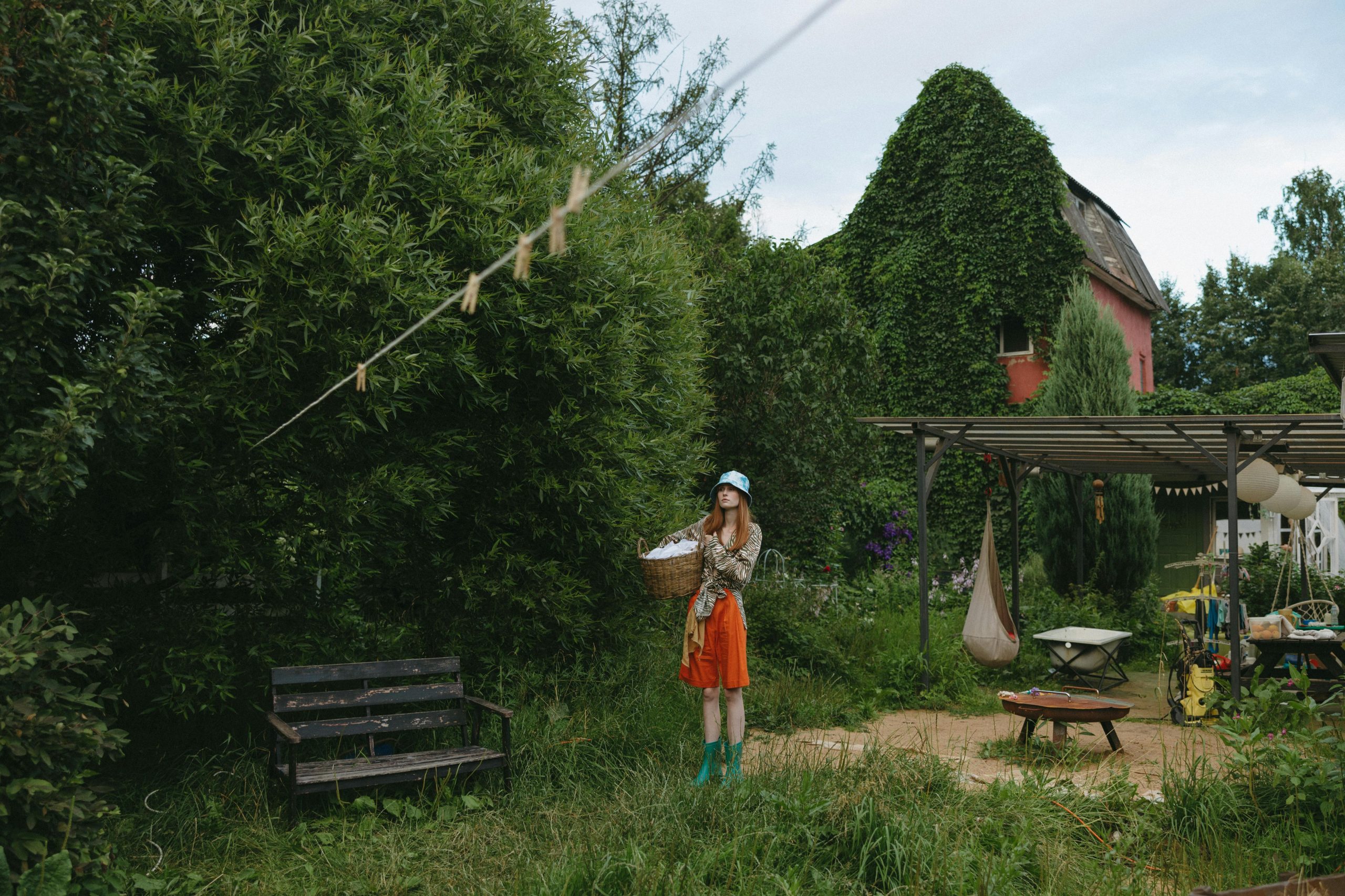 A woman stands in a yard holding a basket, surrounded by greenery and flowers